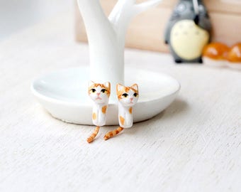May include: A pair of white and orange cat earrings. The cats are facing forward with their tails curled behind them.