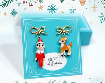 May include: A pair of Christmas-themed earrings featuring a red elf and a brown reindeer. The earrings are on a turquoise blue card with the text "Cat Brothers" printed on it.