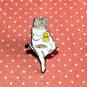 Op de afbeelding: Een emaille pin van een witte en grijze kat, die een bierpul vasthoudt met het woord "BEER". De kat zit met een bord eten ernaast. De pin staat op een rode achtergrond met witte stippen.