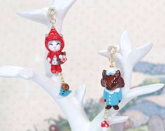 May include: Whimsical dangle earrings featuring a cat in a red hooded cloak and a brown fox in a blue outfit. The earrings are suspended from gold-colored posts with clear gems, and include a snail and mushroom charm.