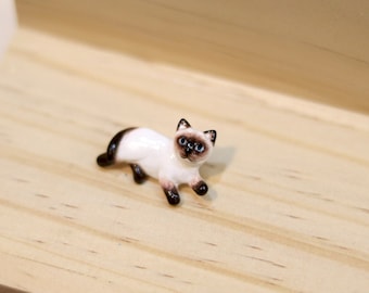May include: A small ceramic figurine of a Siamese cat lying down with its head turned to the side. The cat is white with brown markings on its face and ears.