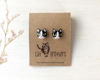 May include: A pair of black and white cat face stud earrings. The earrings are handmade by Cat Brothers. The earrings are on a brown card with the text "Cat Brothers Handmade by Bonnie www.facebook.com/CatBrothersFans"