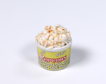 May include: A small yellow and white popcorn container filled with white popcorn kernels. The container has the word "POPCORN" printed on it in red letters.