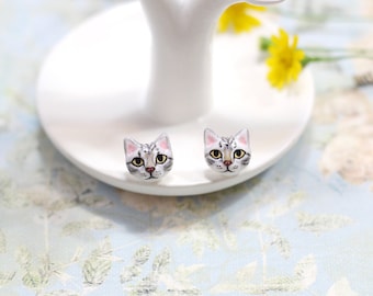 May include: A pair of silver cat face earrings. The cat faces are painted with white, gray, and black fur, and have pink noses and yellow eyes.