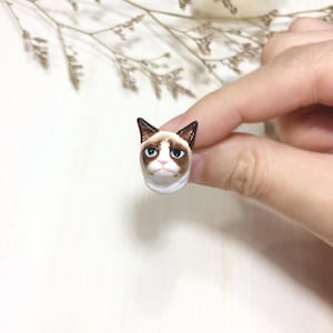 Può includere: Una piccola statuetta in ceramica di un gatto scontroso con pelliccia marrone e segni bianchi. Il gatto ha una faccia bianca con occhi marroni e un naso rosa.