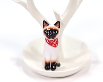 May include: A ceramic figurine of a Siamese cat wearing a red bandana. The cat is sitting upright with its tail curled around its legs. The figurine is on a white ceramic ring holder.