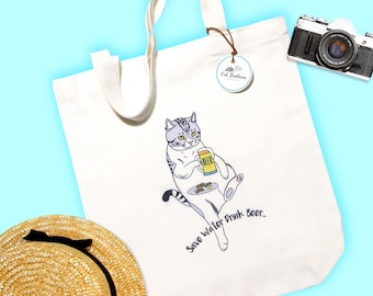 May include: A white canvas tote bag with a cartoon illustration of a cat holding a beer and the text "Save Water Drink Beer." The bag has a brown leather handle and a small white tag with the text "Cat Stickers" on it.