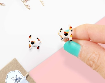 May include: A pair of small, ceramic cat earrings. The cats are painted in a calico pattern with white, orange, and black markings. The earrings are a cute and whimsical accessory for cat lovers.