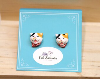 May include: A pair of cat face earrings on a blue display card. The earrings are made of white, orange, and black ceramic and feature a red bandana with a white pattern. The card has the text "Cat Brothers" in white letters on a white oval.