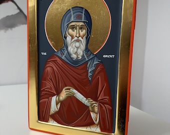 May include: A framed icon of Saint Anthony the Great, painted in a traditional style. The icon features a gold background with a blue halo around the saint's head. The saint is depicted with a white beard and a red robe, holding a scroll in his hands. The text "SAINT ANTHONY THE GREAT" is written in gold lettering at the top of the frame.