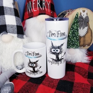 May include: Two white mugs with black cat illustrations and the text "I'm Fine Everything is Fine". One mug is a standard size and the other is a taller travel mug. The mugs are sitting on a red and black plaid blanket.