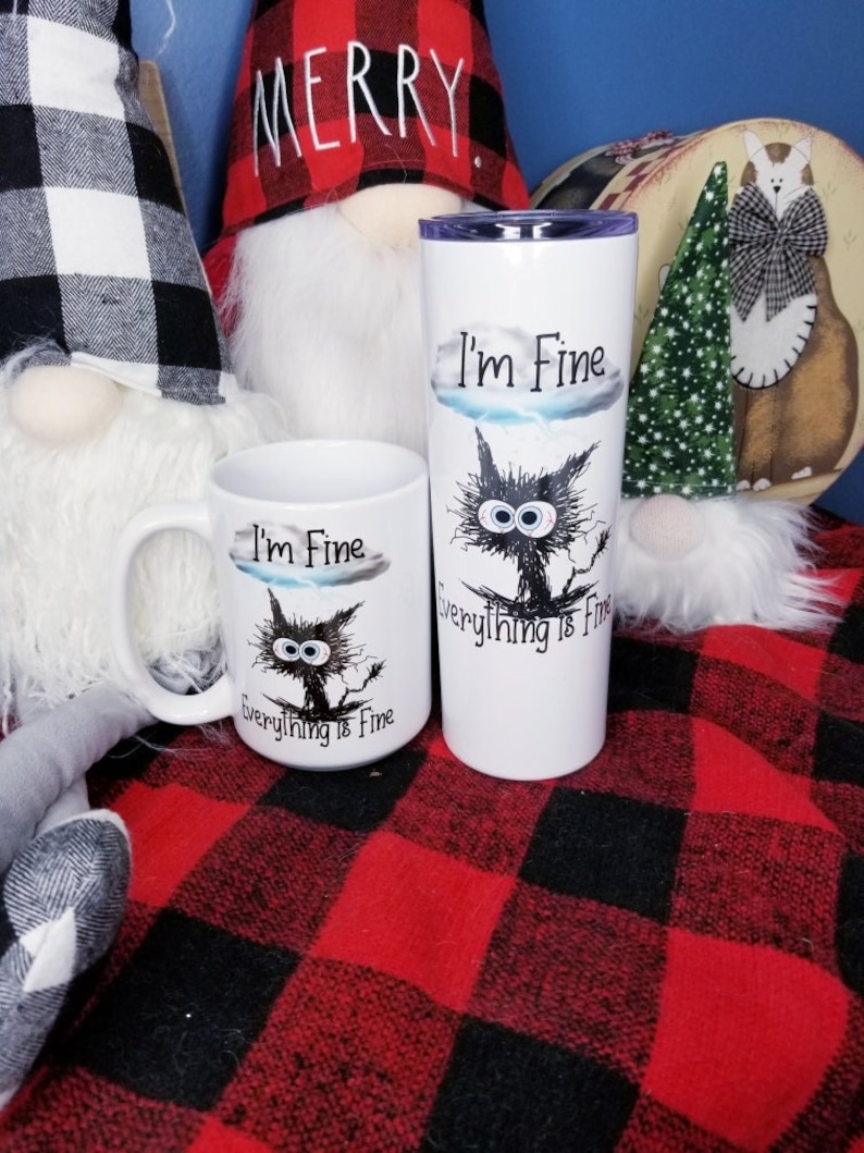 May include: Two white mugs with black cat illustrations and the text "I'm Fine Everything is Fine". One mug is a standard size and the other is a taller travel mug. The mugs are sitting on a red and black plaid blanket.