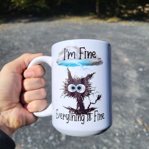 May include: White ceramic mug with a black and white illustration of a cat with a storm cloud above it. The text "I'm Fine" is above the cloud and "Everything is Fine" is below the cat.