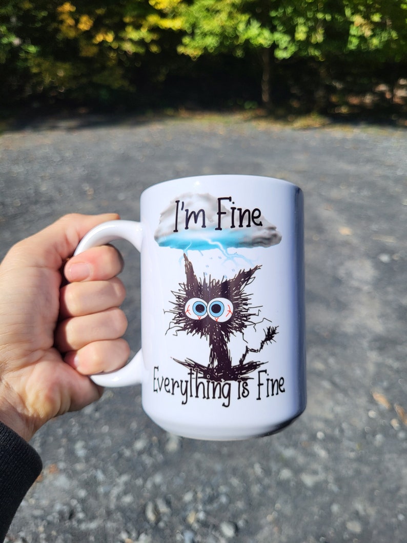 May include: White ceramic mug with a black and white illustration of a cat with a storm cloud above it. The text "I'm Fine" is above the cloud and "Everything is Fine" is below the cat.