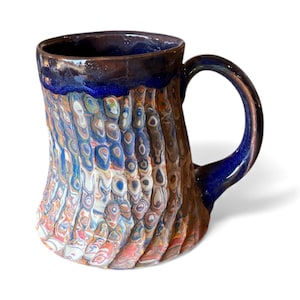 Pode incluir: Caneca de cerâmica artesanal com uma superfície texturizada única. A caneca apresenta uma vibrante mistura de cores, incluindo azul, laranja e branco, com uma borda e alça azul escura. Projetada para bebidas quentes.