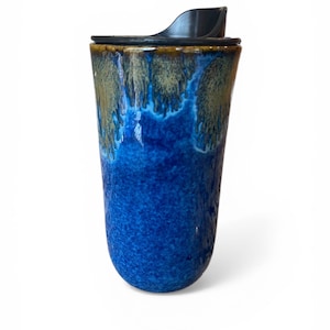 Może przedstawiać: Niebieski ceramiczny kubek podróżny z czarną pokrywką. Kubek ma błyszczące wykończenie z gradientem kolorów, przechodzącym od ciemnoniebieskiego na dole do jaśniejszego niebieskiego i brązowego na górze. Pokrywka ma otwarcie typu flip-top.
