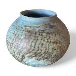 May include: A round ceramic vase with a wide opening. The vase has a textured surface with a pattern of brown and tan lines over a light blue-green base. The rim is a lighter shade of blue.