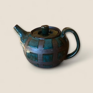 Può includere: Teiera in ceramica con smalto verde scuro e motivo a griglia in una tonalità più scura. Presenta un manico curvo, un beccuccio e un coperchio con un piccolo pomello. Il design è rustico e fatto a mano.