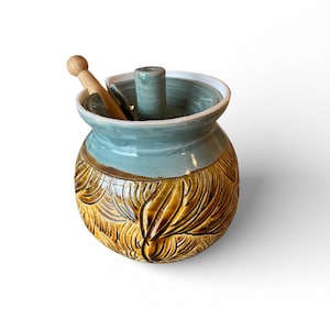 May include: A handcrafted ceramic butter dish with a light blue interior and rim, and a textured, leaf-like pattern in shades of brown and yellow. Includes a small wooden butter knife and a cylindrical holder.