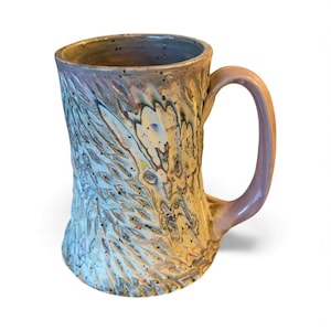 Pode incluir: Uma caneca de cerâmica artesanal com um design texturizado e orgânico. A caneca apresenta uma alça roxa clara e um corpo com uma mistura de cores, incluindo bege, azul e toques de vermelho e amarelo. O exterior da caneca tem um padrão esculpido único.