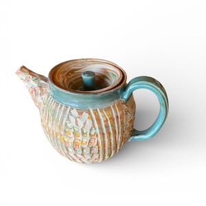 Può includere: Una teiera in ceramica con un corpo strutturato multicolore e un manico e bordo turchese. Il coperchio presenta un motivo a vortice. La teiera ha un beccuccio ed è progettata per preparare il tè.