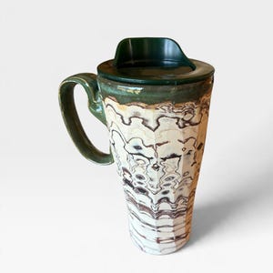 May include: A tall ceramic travel mug with a dark green lid and handle. The body features a faceted, textured surface with a marbled pattern of cream, white, and dark brown wavy lines that wrap around the exterior of the vessel.