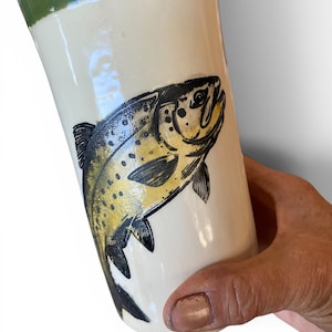 May include: A cylindrical ceramic tumbler with a glossy off-white finish and a dark green band around the top rim. The body features a detailed black and yellow illustration of a trout fish with visible scales, fins, and spots in a side profile.