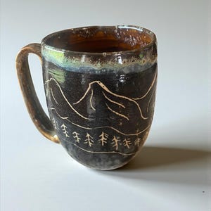 Könnte beinhalten: Handgefertigte Keramik-Tasse mit dunkler, strukturierter Oberfläche. Die Tasse zeigt ein Berg- und Baumdesign, das in Weiß geätzt ist. Der Rand hat eine braune und grüne Glasur, und der Griff ist in einer ähnlichen braunen Farbe gehalten.