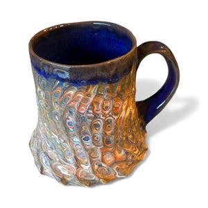 Peut inclure: Une tasse en céramique faite à la main avec un extérieur texturé unique. La tasse présente un motif tourbillonnant de couleurs, notamment le bleu, l'orange et le blanc. L'intérieur et la poignée sont bleu foncé, créant un contraste saisissant. Un objet fonctionnel et décoratif.