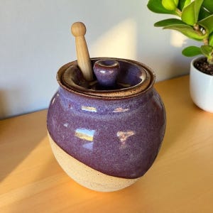 Puede incluir: Un tarro de cerámica hecho a mano con tapa y un cucharón de miel de madera. El tarro es de color púrpura oscuro con una base y borde beige. El cucharón de miel está hecho de madera clara. El tarro se utiliza probablemente para guardar miel u otros condimentos.
