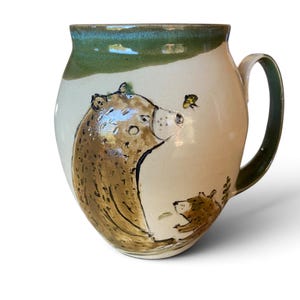 May include: A ceramic mug with a cream-colored base and a green glazed rim. The mug features a hand-painted illustration of a large brown bear looking at a small bee, with a smaller bear cub sitting below. It includes a curved handle.