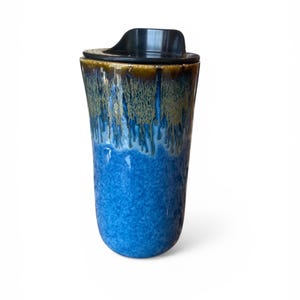 Może przedstawiać: Ceramiczny kubek podróżny z czarną pokrywką. Kubek ma niebieskie i brązowe szkliwione wykończenie. Niebieska glazura pokrywa dolną część kubka, a brązowa glazura znajduje się na górze. Kubek został zaprojektowany tak, aby napoje były gorące lub zimne.