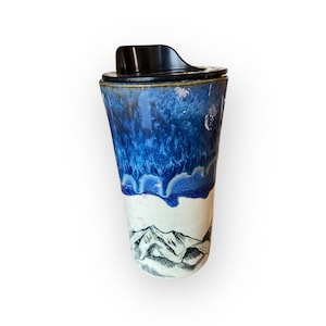 Pode incluir: Uma caneca de viagem de cerâmica com tampa preta. A caneca apresenta um esmalte azul e branco, com um desenho de cordilheira na base. A caneca tem aproximadamente 15 cm de altura.