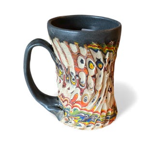 May include: A handcrafted ceramic mug with a distinctive, textured finish. The mug has a black rim and handle, contrasting with the colourful, swirling pattern of white, orange, yellow, and blue. The mug is designed for hot drinks.