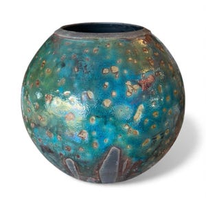 May include: A spherical ceramic vase with a speckled, iridescent finish. The vase has a blend of teal, blue, and green hues with brown and gold accents. The opening is dark, and the overall shape is round.