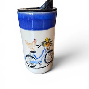 May include: A ceramic travel mug with a glossy white finish and a deep blue glazed rim. The body features a hand-painted illustration of a light blue bicycle with a basket of yellow flowers and a small chicken perched on the seat. Includes a black lid.