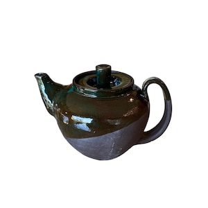 Puede incluir: Una tetera de cerámica con la parte superior esmaltada en verde oscuro y la parte inferior gris mate. La tetera tiene un asa curva y un pico. La tapa tiene un pequeño pomo cilíndrico. La tetera está diseñada para preparar té.