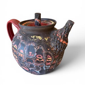 May include: A round ceramic teapot with a dark charcoal base featuring a textured, dimpled surface. The body displays swirling patterns of red, yellow, and blue. It has a solid red handle, a matching lid with a vertical handle, and a textured spout.