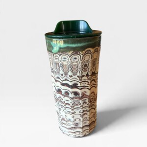 May include: A tall ceramic travel mug featuring a dark green lid and a cylindrical body with a faceted surface. The exterior displays a complex, layered marbled pattern in shades of cream, brown, and dark green, creating a repeating wavy design.