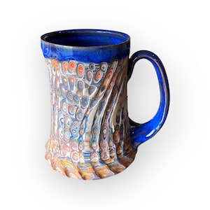 May include: A handcrafted ceramic mug with a blue rim and handle. The body of the mug features a colorful, textured pattern with swirls of orange, white, and blue. The mug has a capacity of approximately 16 ounces.
