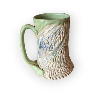 Pode incluir: Caneca de cerâmica artesanal com alça e borda verde claro. O corpo da caneca apresenta um design texturizado e esculpido com detalhes em azul, branco e laranja. A caneca tem um formato único com uma alça confortável.