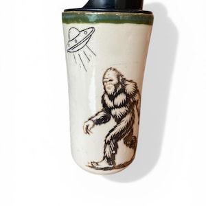 Op de afbeelding: Een witte keramische reismok met een zwarte deksel en een groene rand. De mok heeft een zwarte illustratie van een UFO en een bruine en zwarte illustratie van Bigfoot. De mok is ontworpen voor warme of koude dranken.