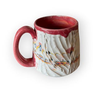 Puede incluir: Una taza de cerámica hecha a mano con un exterior blanco texturizado y un interior y asa rojos. La taza presenta un diseño tallado con acentos coloridos de amarillo, azul y naranja. El borde también es rojo.