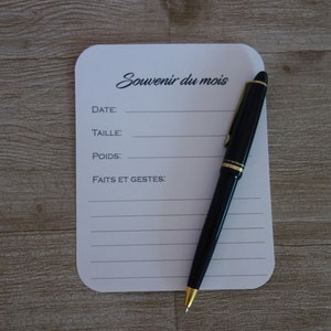 May include: A white card with the text "Souvenir du mois" and lines for writing down the date, size, weight, and events of the month. A black pen with a gold trim is lying on the card.