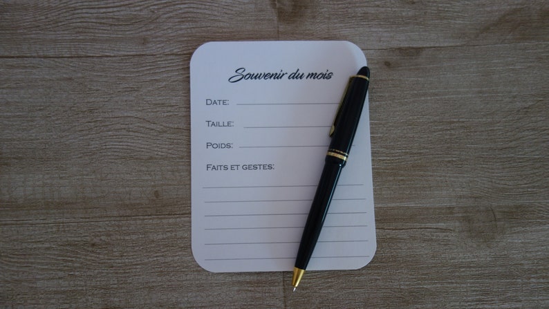 May include: A white card with the text "Souvenir du mois" and lines for writing down the date, size, weight, and events of the month. A black pen with a gold trim is lying on the card.