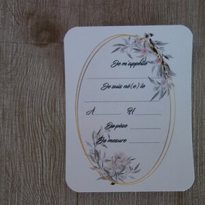 May include: A white card with a gold oval frame decorated with floral illustrations. The card has the text "Je m'appelle", "Je suis n&eacute;(e) le", "d", "H", "De p&egrave;se", and "De mesure".