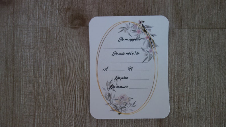 May include: A white card with a gold oval frame decorated with floral illustrations. The card has the text "Je m'appelle", "Je suis n&eacute;(e) le", "d", "H", "De p&egrave;se", and "De mesure".