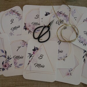 May include: A set of twelve cards with floral designs and gold accents. Each card features a number from 1 to 12, with the word "Mois" (French for "month") written below. The cards are designed for a baby's first year.