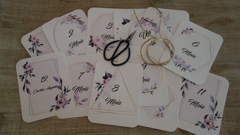 May include: A set of twelve cards with floral designs and gold accents. Each card features a number from 1 to 12, with the word "Mois" (French for "month") written below. The cards are designed for a baby's first year.