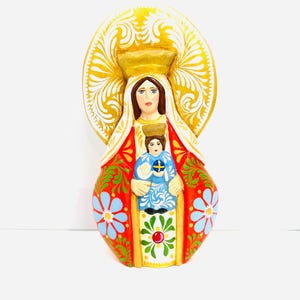 Może przedstawiać: Ręcznie malowana drewniana figurka Matki Boskiej z Dzieciątkiem Jezus. Matka Boska ma na sobie czerwoną szatę z niebieskimi i białymi wzorami kwiatowymi. Dzieciątko Jezus ma na sobie niebieską szatę z złotą aureolą. Figurka ma złotą aureolę wokół głowy Matki Boskiej.